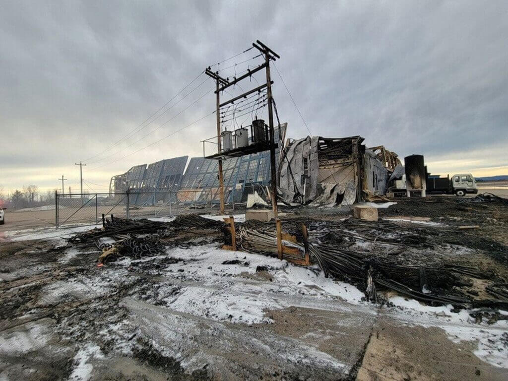 Burned industrial building with charred debris and power lines against a cloudy sky.