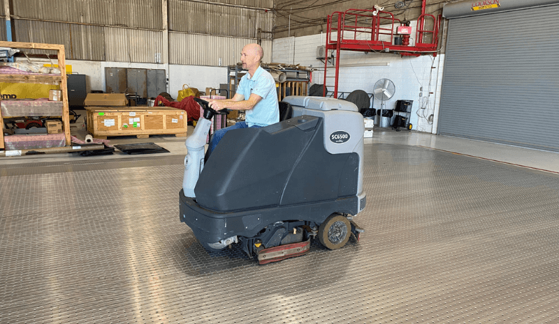 Man operating a floor scrubber in an industrial warehouse setting.