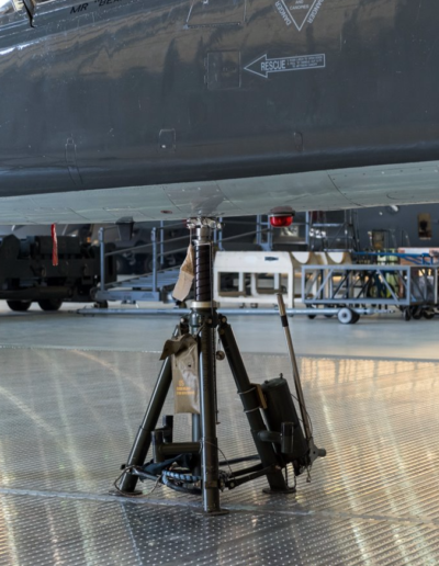 Aircraft in hangar, supported by a maintenance tripod jack on a reflective metal floor.