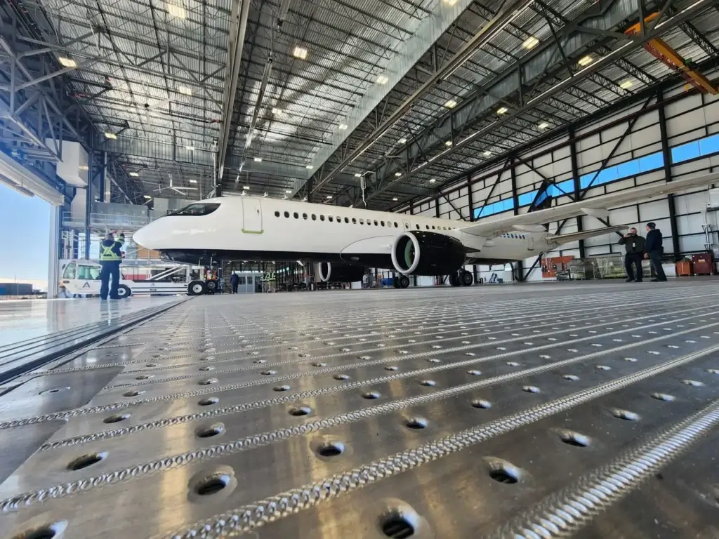 Workers guide a large aircraft inside a spacious hangar with a textured metal floor.