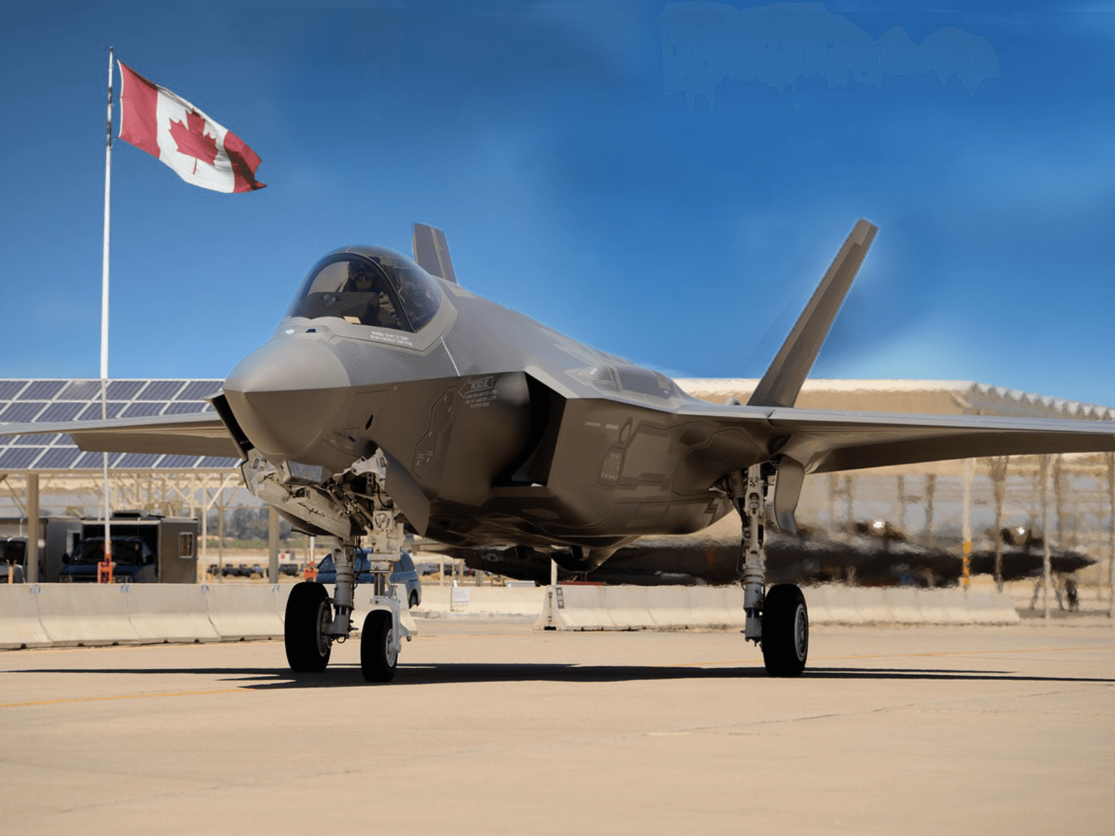 Jet fighter taxiing on runway with Canadian flag in the background under a clear blue sky.