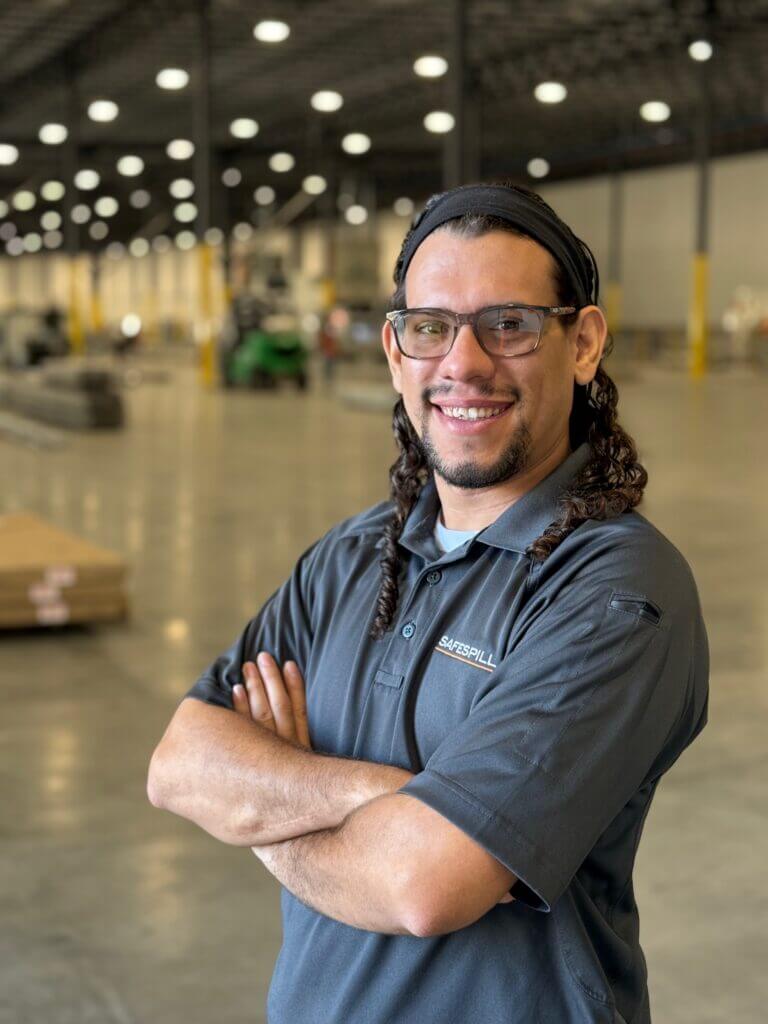 Man smiling with arms crossed in a spacious warehouse, wearing a Safespill polo and glasses.
