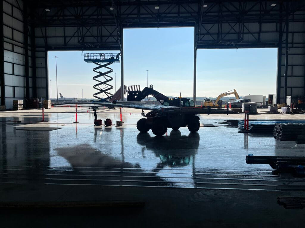 Construction equipment in a large hangar with open doors, silhouette against bright sky.