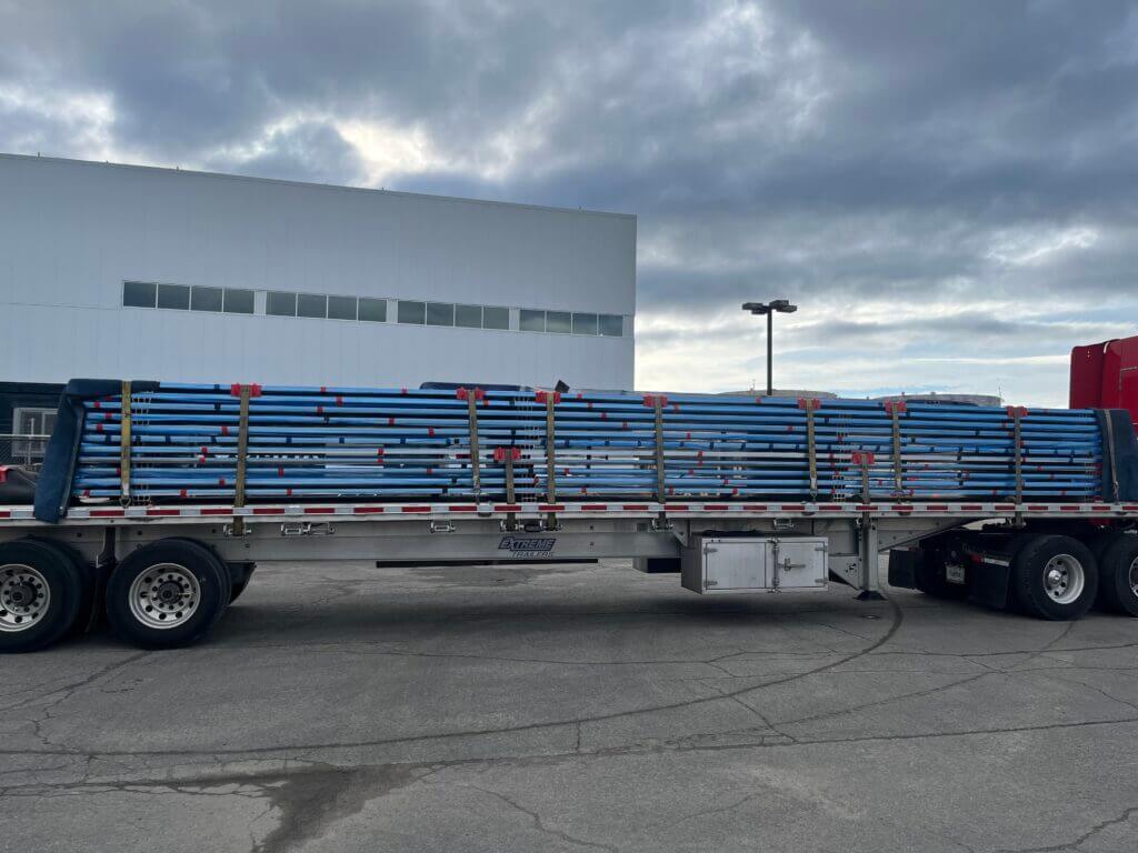 Flatbed trailer loaded with stacked blue pipes, secured with straps, parked in an industrial area under a cloudy sky.