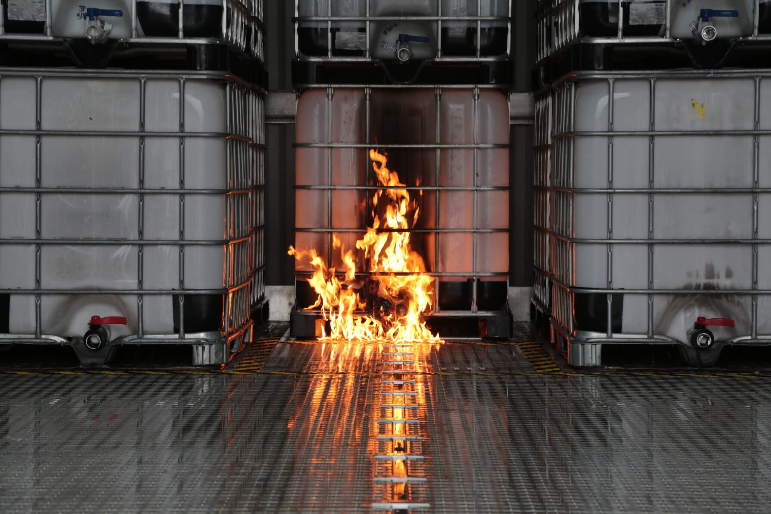 Fire burning on a metal floor between large storage containers.