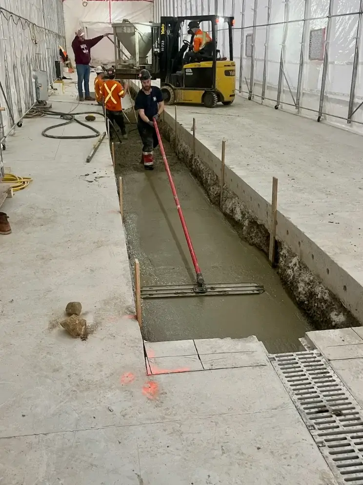 Workers smoothing freshly poured concrete in a long trench inside a construction site.
