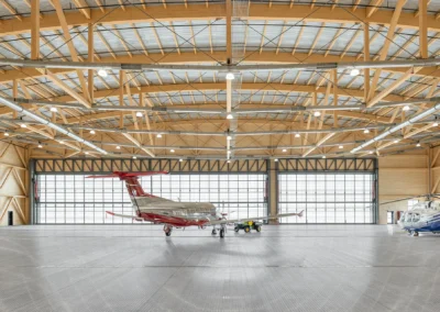 Airplane and helicopter parked inside a spacious, sunlit hangar with wooden beams and large windows.