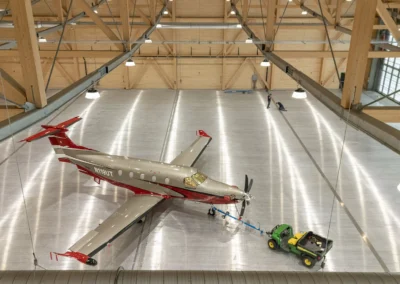 Small aircraft being towed by a green tractor inside a spacious, wooden-frame hangar.