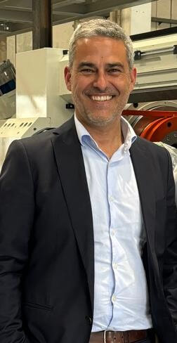Smiling man in a suit standing indoors, industrial equipment in the background.