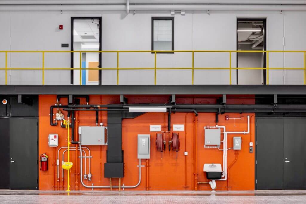 Industrial wall with equipment, yellow railing, and open doors above. Pipes, fire extinguisher, and safety wash station visib