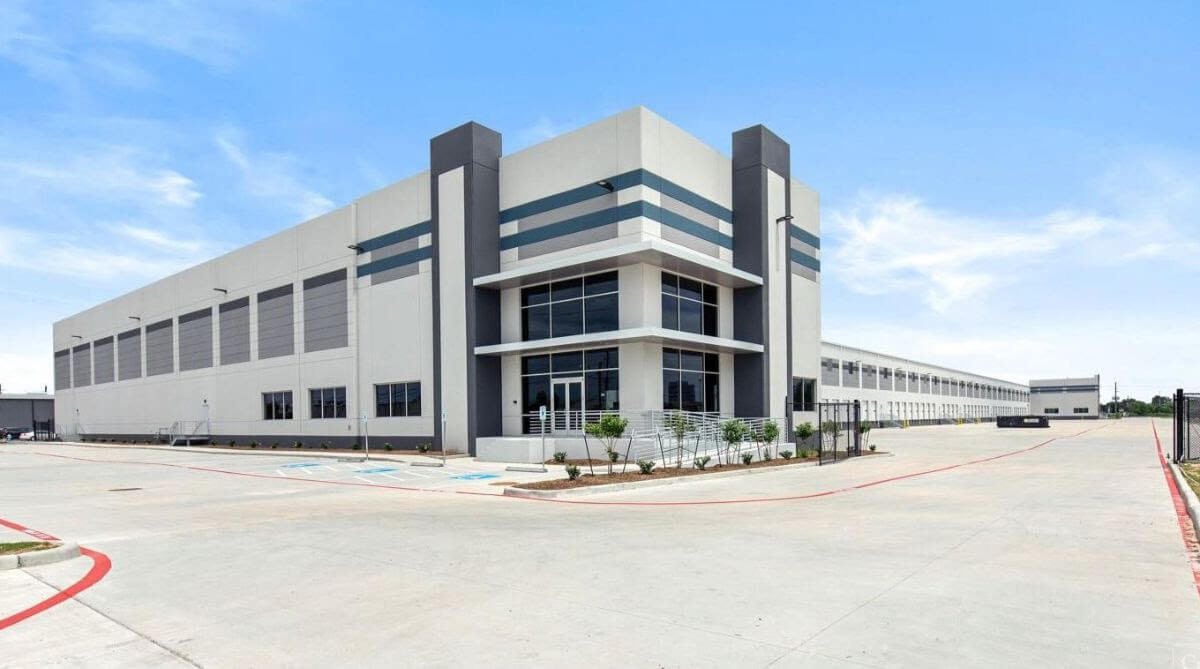 Modern warehouse building with large windows and a spacious parking lot under a clear blue sky.